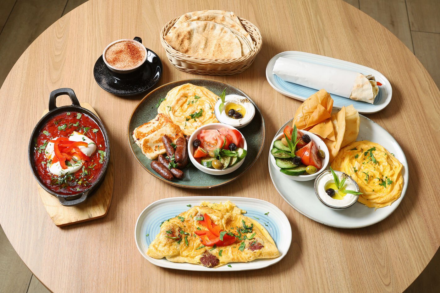 A breakfast table with shakshuka, The Lebanese spread, morning mezze, an omelette, a sausage and egg wrap and a cappuccino